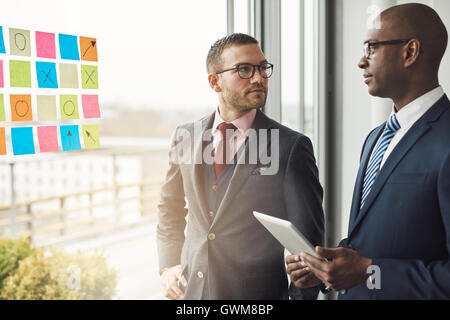 Caucasian and African businessman in a meeting standing in front of colorful memos on the glass of a window in the office lookin Stock Photo