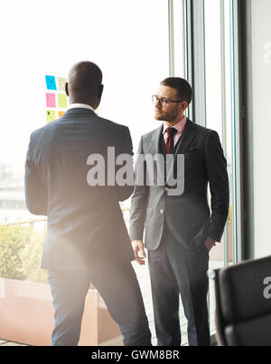 Serious young businessman wearing glasses standing in front of a bright window overlooking a patio talking to an African colleag Stock Photo