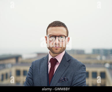 Determined intense young businessman wearing glasses standing on an outdoor patio of a high rise building staring at the camera Stock Photo