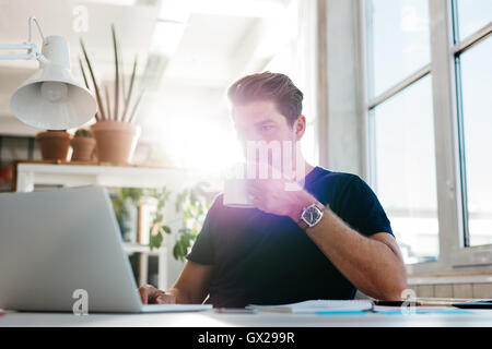 Young businessman drinking coffee and looking at laptop screen at workplace. Male executive working at his desk and drinking cof Stock Photo