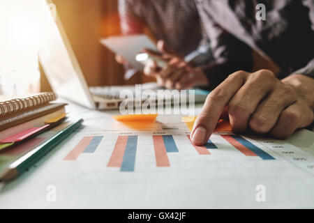 business documents on office table with tablet, smart phone and laptop and two colleagues discussing data in morning light Stock Photo