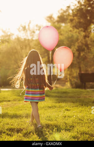 Little girl with balloons walks in the park outdoors. Man is unrecognizable. Shooting from a back. Stock Photo