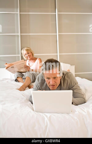 Couple on bed reading newspaper and using laptop Stock Photo