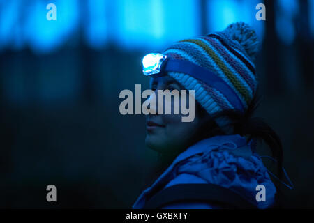 Female hiker wearing head torch looking over her shoulder from forest at night Stock Photo