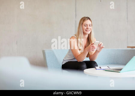 Young female student sitting on study space sofa reading smartphone texts at higher education college Stock Photo