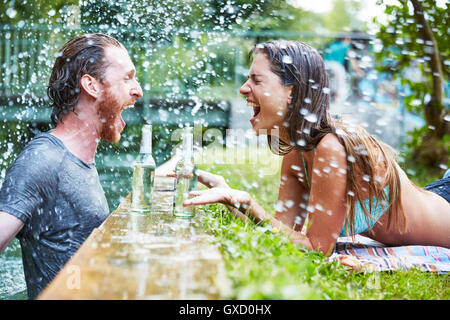 Young man in river, young woman lying on grass at edge of river, man splashing woman with water Stock Photo