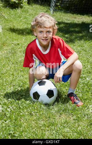 Portrait of boy wearing soccer uniform crouching with soccer ball in garden Stock Photo