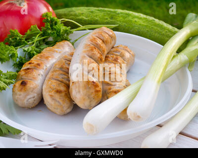 Grilled Munich sausages on the disposable white plastic plate arranged with tomato, cucumber green onion and parsley Stock Photo