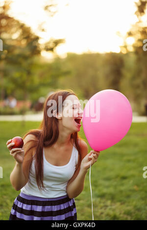 Young attractive woman biting a balloon. She is holding a red apple. Girl plays the fool. Stock Photo