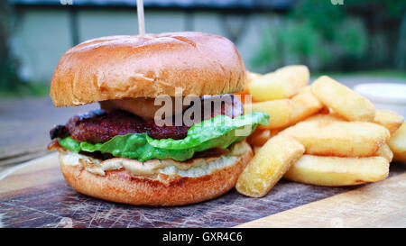 Close up of meat burger with green lettuce, relish in a wheat bun offered on wooden board, accompanied by chunky potato fries Stock Photo