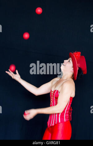 Smiling female juggler wearing a red top hat and red basque with three balls in the air and one in the hand Stock Photo