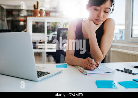 Shot of asian businesswoman writing notes on notepad. Female executive sitting at her desk with laptop taking notes. Stock Photo