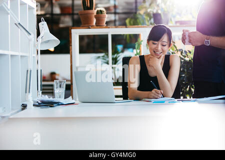 Shot of smiling asian businesswoman writing notes on notepad with colleague standing by. Female executive working at her desk wi