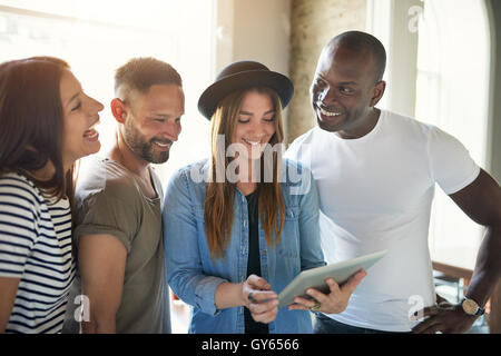 Male and female friends smile at computer tablet while standing together by large windows as sun pours in Stock Photo