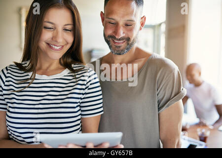 Smiling couple hold computer tablet between them while friends sit at nearby table Stock Photo