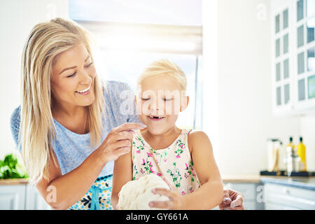 Cute happy little girl missing her front teeth learning to bake with her mother in the kitchen as she helps with kneading the do Stock Photo