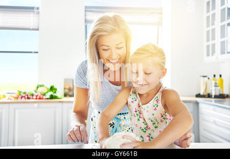 Adorable little girl kneading the dough watched over by her loving mother as she learns to bake, high key sun flare background Stock Photo