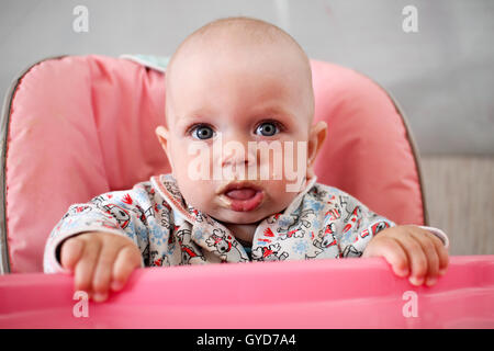 Beautiful baby eats porridge from mom's hand. He is sitting on a pink children' chair. Stock Photo