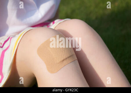 A girl sitting on the grass in a stripy skirt with a big plaster on her knee.  Close up. Stock Photo