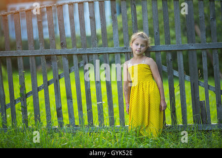 Little girl in yellow dress near the wooden fence in the village. Stock Photo