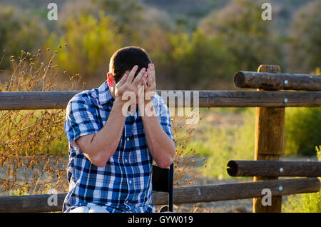 Man upset with head in his hands sitting outdoors with a vibrant field in the background. Stock Photo
