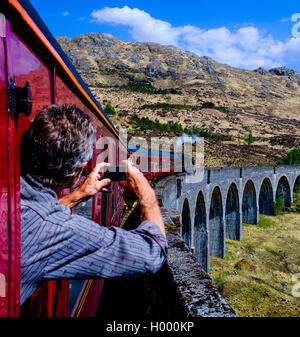 Man leans out the window taking a picture of historic train 'The Jacobite' Steam Train driving over the Glenfinnan Viaduct Stock Photo