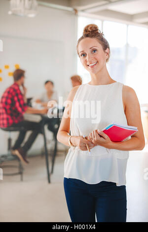 Portrait of happy young business woman with coworkers meeting in background. Female executive in casuals standing in modern offi Stock Photo
