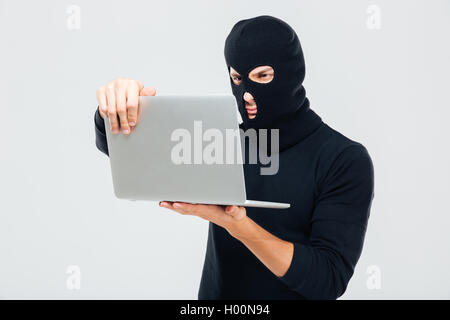 Criminal young man in balaclava standing and using laptop Stock Photo