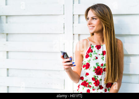 Beautiful young woman using phone while leaning against a white wall during a bright summer day Stock Photo