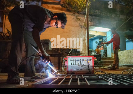 KAMALAPURAM, INDIA - 02 FEBRUARY 2015: Indian worker welding heavy metal parts on street in dusk Stock Photo