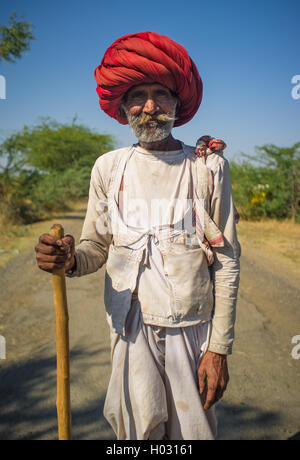 GODWAR REGION, INDIA - 14 FEBRUARY 2015: Elderly Rabari tribesman with big red turban and cane stands on road. Rabari or Rewari  Stock Photo