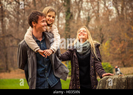 Daughter rides father on piggyback with mother next to them in park on an autumn day. Stock Photo