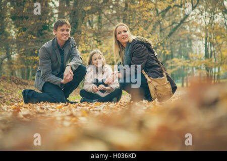 Cheerful family of three kneel on park ground covered with leaves on an autumn day. Stock Photo