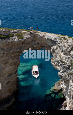 An excursion boat under the Trypitos Arch and tourists walking on top of the limestone arch over the sea on Paxos, The Ionian Is Stock Photo