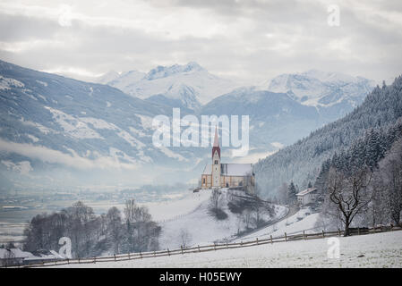 St. Pankraz church in an alpine landscape in the snow surrounded by clouds and fog on a hill in Zillertal valley, Austria. Stock Photo