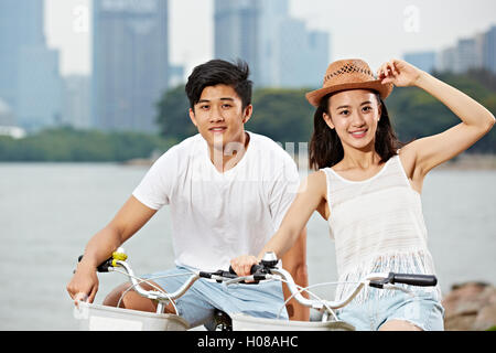 young asian couple riding bike in urban park, looking at camera smiling Stock Photo