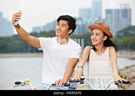 young asian couple taking a selfie while riding bike in urban park Stock Photo