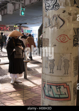 Takuhatsu. A Buddhist monk in traditional robes, begging for alms, next to graffitti covered wall, Ueno Station, Tokyo, Japan Stock Photo