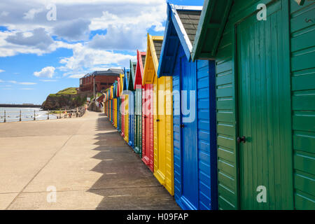 Row of colourful beach huts and their shadows, distant surfers in sea, West Cliff Beach, Whitby, North Yorkshire, England, UK Stock Photo