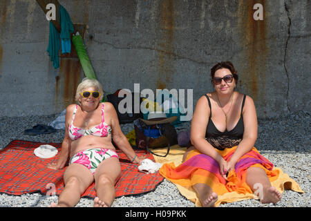 Two plump women, senior smiling elderly blonde and mature brunette are sitting on pebble beach on concrete retaining wall backgr Stock Photo