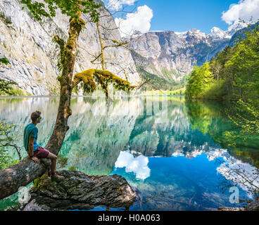 Young man sitting on tree above water, looking into distance, reflection in lake, Obersee, Salet am Königssee Stock Photo