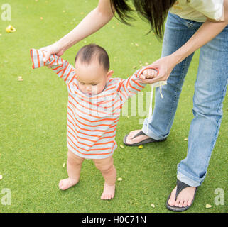 Asian mother holding baby hand to walk training  on the green glass field Stock Photo