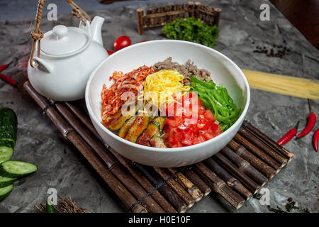 Korean noodle with kimchi, tomatoes, chili in white bowl on bamboo tray with tea pot Stock Photo