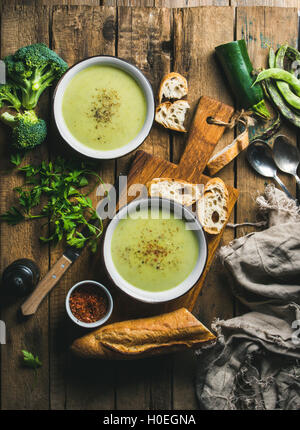 Two bowls of homemade pea, broccoli and zucchini cream soup served with fresh baguette and vegetables on wooden board over rusti Stock Photo