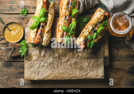 Glass of wheat unfiltered beer and grilled sausage dogs in baguette on baking paper with mustard, caramelised onion and herbs on Stock Photo