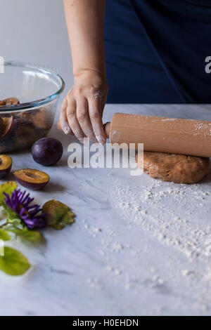 Woman rolling out dough on a floured white marble table, ingredients for baking plum galette Stock Photo