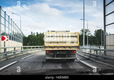 Big industrial tipper truck goes on asphalt road, rear view from other car cabin Stock Photo