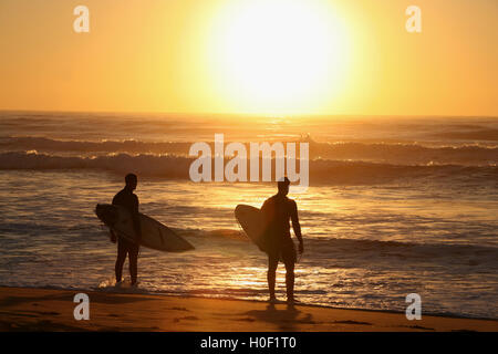Sunset silhouette of two surfers standing on the beach with surfboards under their arms looking out to see Stock Photo