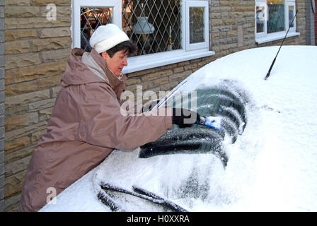 Women in warm winter clothing with scraper removing frozen snow from Ford Ka car windshield parked in home driveway England UK Stock Photo