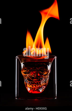 Skull shape glass with flames on the top. Single object on black background. Fire as the shape of tail and horns. Dark and moody image of the drink. Stock Photo